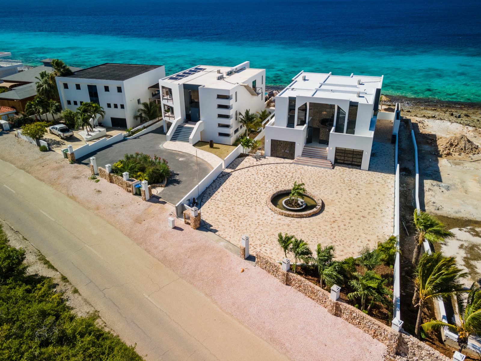 Aerial view of a modern luxury oceanfront villa in Bonaire, featuring a white contemporary facade, a grand stone driveway, and direct access to the turquoise Caribbean water.